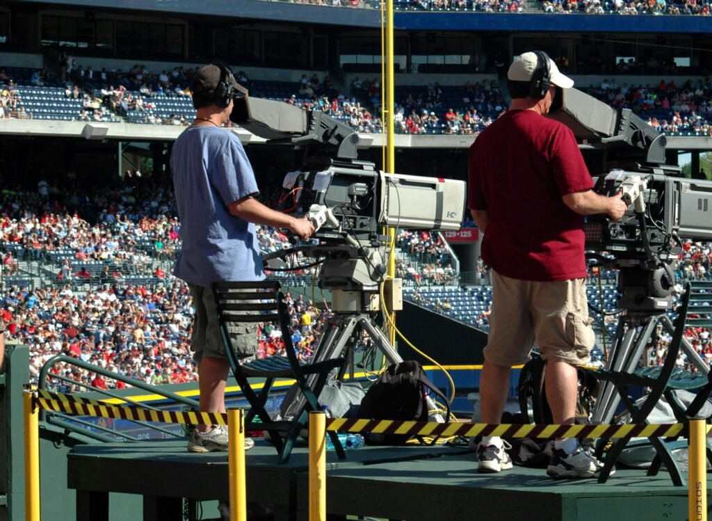 Camera operators capturing a live sports event at a crowded outdoor stadium filled with spectators.