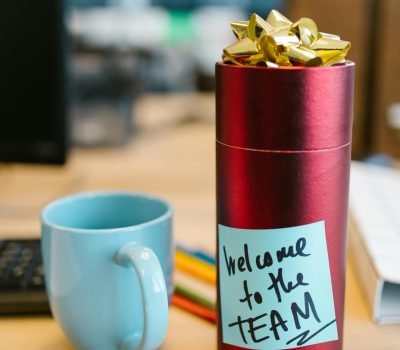 Red gift on desk with sticky note 'Welcome to the TEAM' beside blue mug, ideal for workplace imagery.